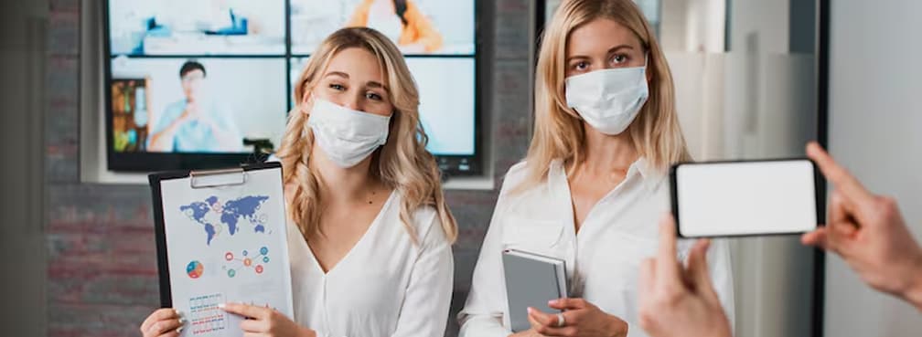Two women wearing face masks hold data charts while being filmed or photographed by a smartphone.
