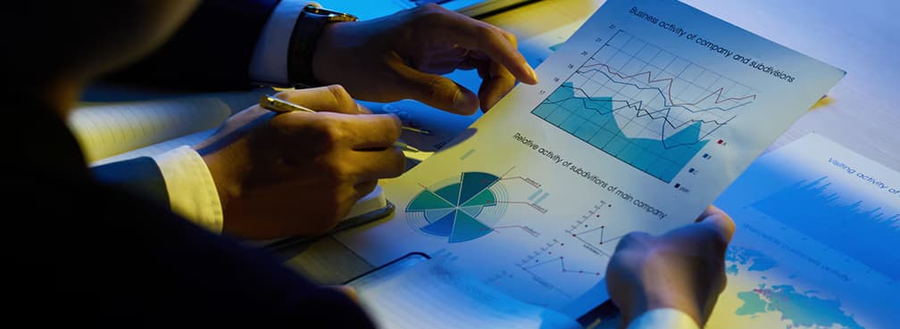Hands holding and pointing at printed business performance charts during a late-night meeting.