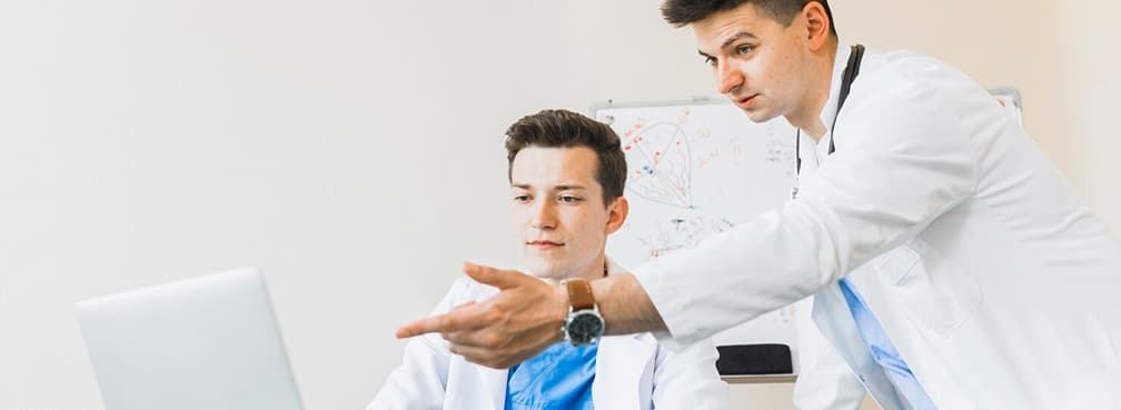Young physician gesturing toward a laptop screen while training a medical colleague.