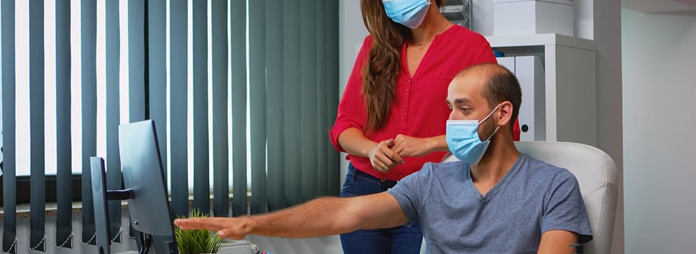 Professional teammates wearing masks while discussing a project at a desktop workstation.