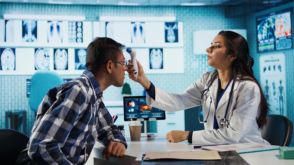 A healthcare professional measuring a man's temperature at a clinic desk consultation.