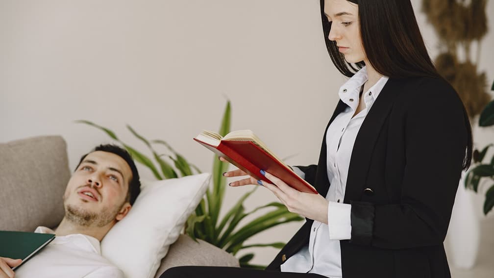 A female counselor reads from a book while a man rests on a couch during a therapy appointment.